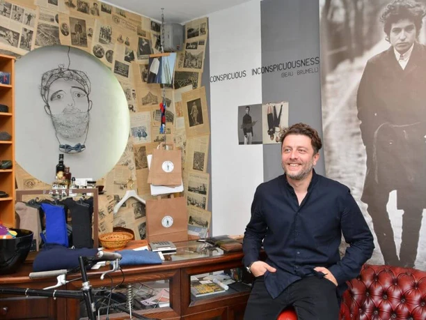 Greenwich Man Time Das Bild zeigt den Ladenbesitzer Evgenij Leschiner von Greenwich Man Time in Köln, der entspannt in einem gemütlichen, individuell gestalteten Raum sitzt. Hinter ihm sind eine Vielzahl von antiken Gegenständen und Zeitungen an der Wand zu sehen, die dem Raum einen Vintage-Charme verleihen.