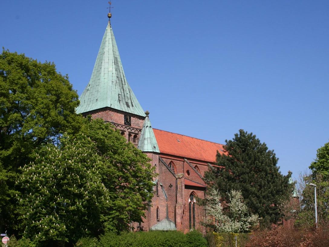 Rote Backsteinkirche mit grünem Kirchturmdach, umgeben von grünen Bäumen unter klarem blauen Himmel.