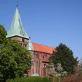 Rote Backsteinkirche mit grünem Kirchturmdach, umgeben von grünen Bäumen unter klarem blauen Himmel.