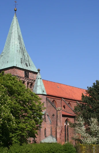 Rote Backsteinkirche mit grünem Kirchturmdach, umgeben von grünen Bäumen unter klarem blauen Himmel.