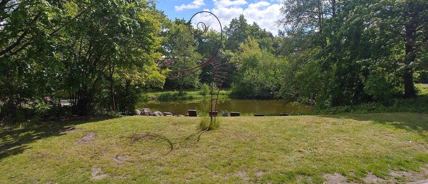 Füllhorn oder Figura Skulptur in einem grünen Park vor einem Teich, umgeben von Bäumen und blauem Himmel mit Wolken.
