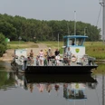 Fähre überquert einen ruhigen Fluss, beladen mit Radfahrern, umgeben von grüner Landschaft.