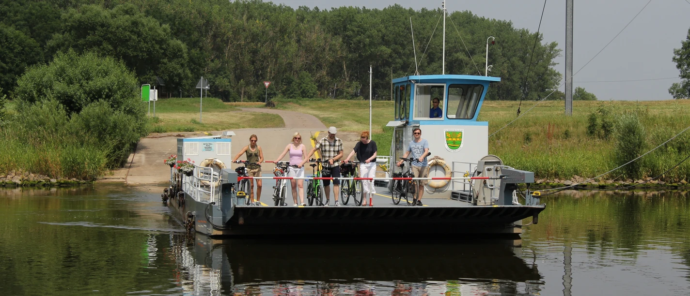 Fähre überquert einen ruhigen Fluss, beladen mit Radfahrern, umgeben von grüner Landschaft.
