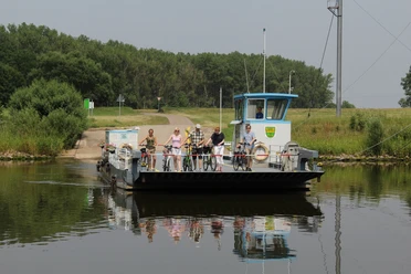 Fähre Schweringen Fähre überquert einen ruhigen Fluss, beladen mit Radfahrern, umgeben von grüner Landschaft.