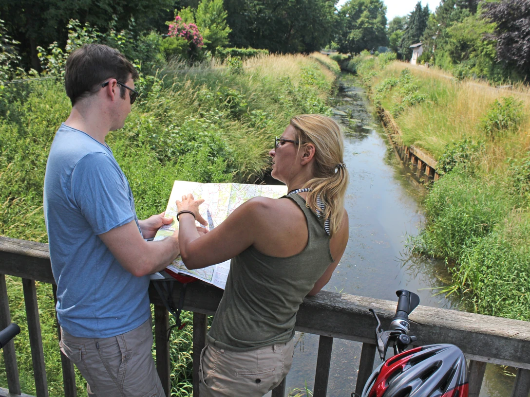 Zwei Radfahrer stehen auf einer Brücke und betrachten eine Karte. Ein Fluss fließt darunter.