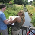 Zwei Radfahrer stehen auf einer Brücke und betrachten eine Karte. Ein Fluss fließt darunter.
