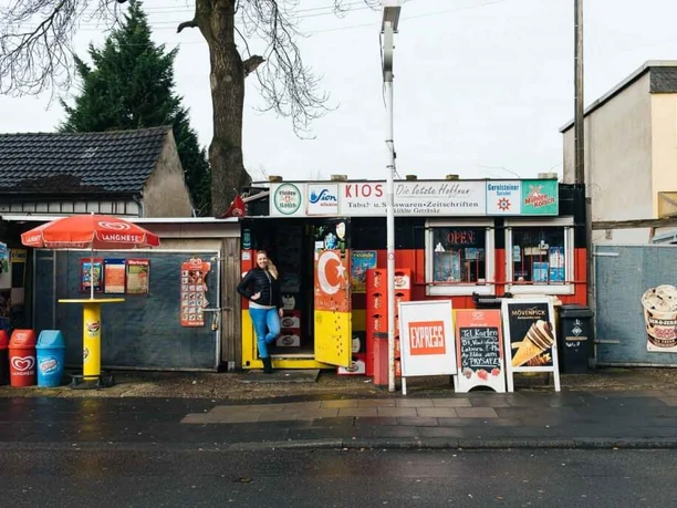 Kiosk Ein traditionelles, buntes Kiosk in Köln, flankiert von Fahnen und Werbung. Asphaltierte Straße im Vordergrund.