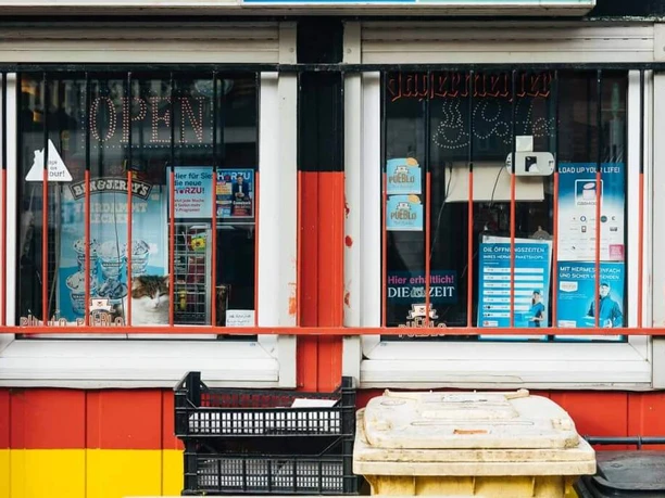 Kiosk Ein typisches "Büdchen" in Köln. Das Kioskfenster ist mit verschiedenen Plakaten und Hinweisschildern dekoriert. Die Fenster sind eingefasst mit einer rot-gelben Farbkombination. Eine beleuchtete "Open"-Anzeige signalisiert, dass der Laden geöffnet ist. Davor sind einige Kisten und eine gelbe Plastiktonne zu erkennen.