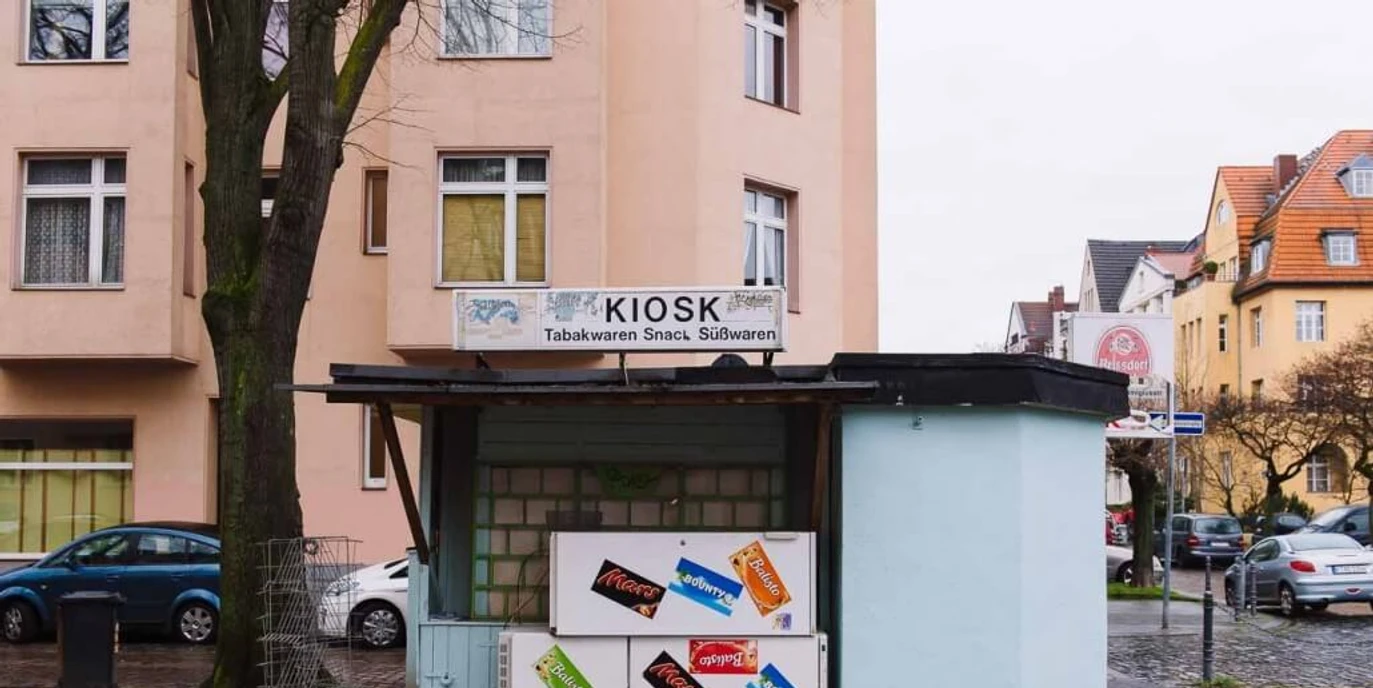 Kiosk at Nikolausplatz Kleiner Kiosk auf einer ruhigen Straßenecke, umgeben von Wohngebäuden, mit Werbetafeln für Snacks.Small kiosk on a quiet street corner surrounded by residential buildings, featuring advertising boards for snacks.