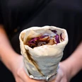 Oruc Kebap Eine Person hält eine Dönerrolle, gefüllt mit roten Kohlstreifen und weiteren Zutaten, umwickelt in Alufolie. A person is holding a kebab roll filled with red cabbage strips and other ingredients, wrapped in aluminium foil.