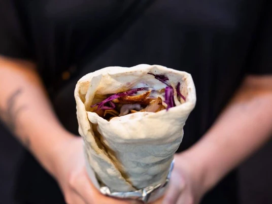 Oruc Kebap Eine Person hält eine Dönerrolle, gefüllt mit roten Kohlstreifen und weiteren Zutaten, umwickelt in Alufolie. A person is holding a kebab roll filled with red cabbage strips and other ingredients, wrapped in aluminium foil.