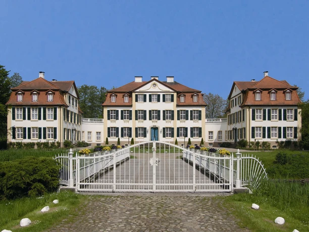 Schloss Hüffe Schloss Hüffe mit imposanter Fassade, weitläufigem Vorplatz und schmiedeeisernem Tor vor blauem Himmel.