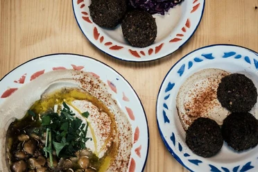 Mashery Hummus Kitchen Auf einem Tisch liegen Teller mit klassischen Mezze. Ein Teller zeigt cremigen Hummus mit Kichererbsen und frischen Kräutern, die in der Mitte angerichtet sind. Die anderen Teller enthalten Falafel-Kugeln, perfekt golden gebraten.
