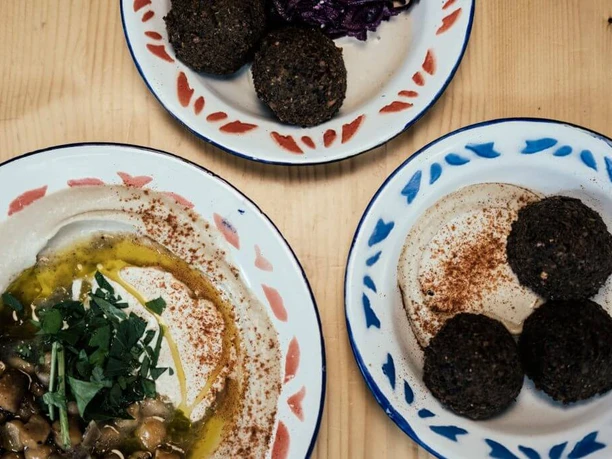 Mashery Hummus Kitchen Auf einem Tisch liegen Teller mit klassischen Mezze. Ein Teller zeigt cremigen Hummus mit Kichererbsen und frischen Kräutern, die in der Mitte angerichtet sind. Die anderen Teller enthalten Falafel-Kugeln, perfekt golden gebraten.