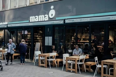 Mama trattoria Menschen sitzen an Holztischen vor einem modernen Restaurant mit dem Schriftzug "mama" und genießen ihre Zeit.