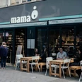 Mama trattoria Menschen sitzen an Holztischen vor einem modernen Restaurant mit dem Schriftzug "mama" und genießen ihre Zeit.People are sitting at wooden tables in front of a modern restaurant with the word ‘mama’ written on it, enjoying their time.