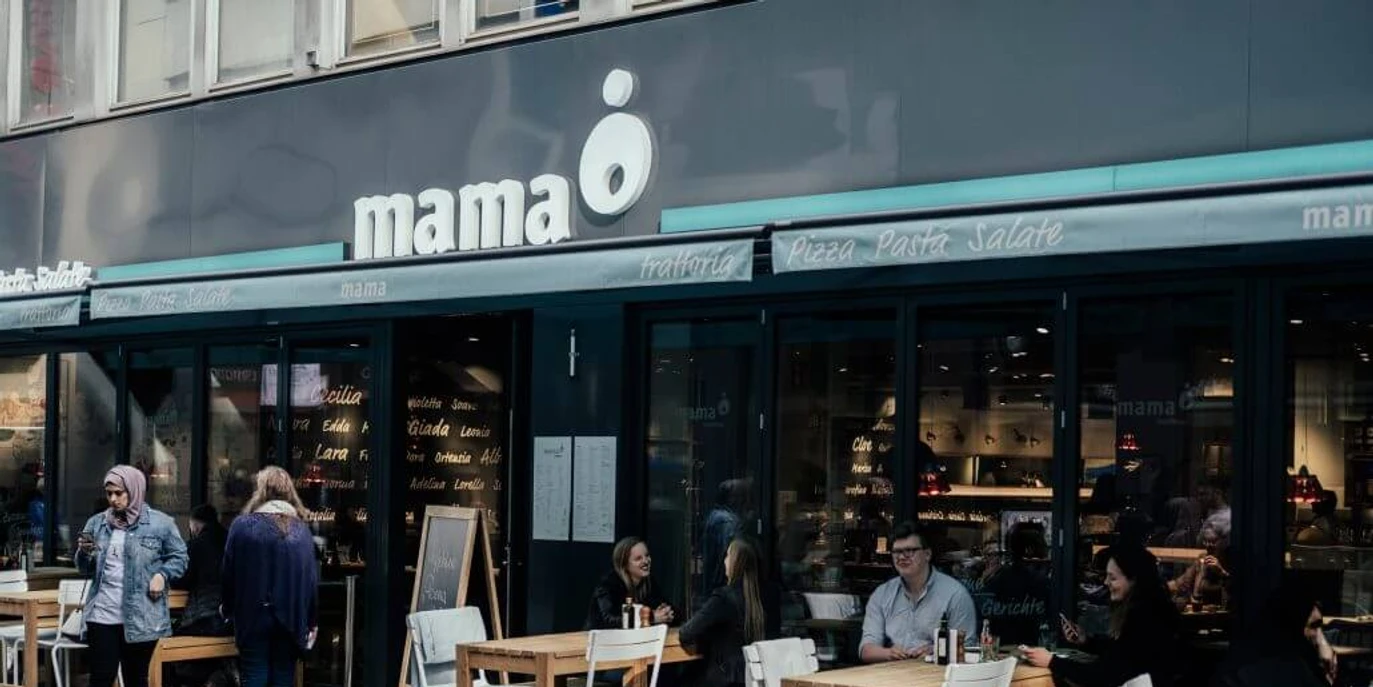 Mama trattoria Menschen sitzen an Holztischen vor einem modernen Restaurant mit dem Schriftzug "mama" und genießen ihre Zeit.People are sitting at wooden tables in front of a modern restaurant with the word ‘mama’ written on it, enjoying their time.