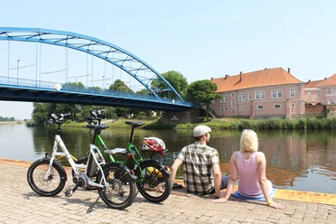 Zwei Personen pausieren an einem Flussufer, neben ihnen stehen Fahrräder, im Hintergrund eine Brücke.