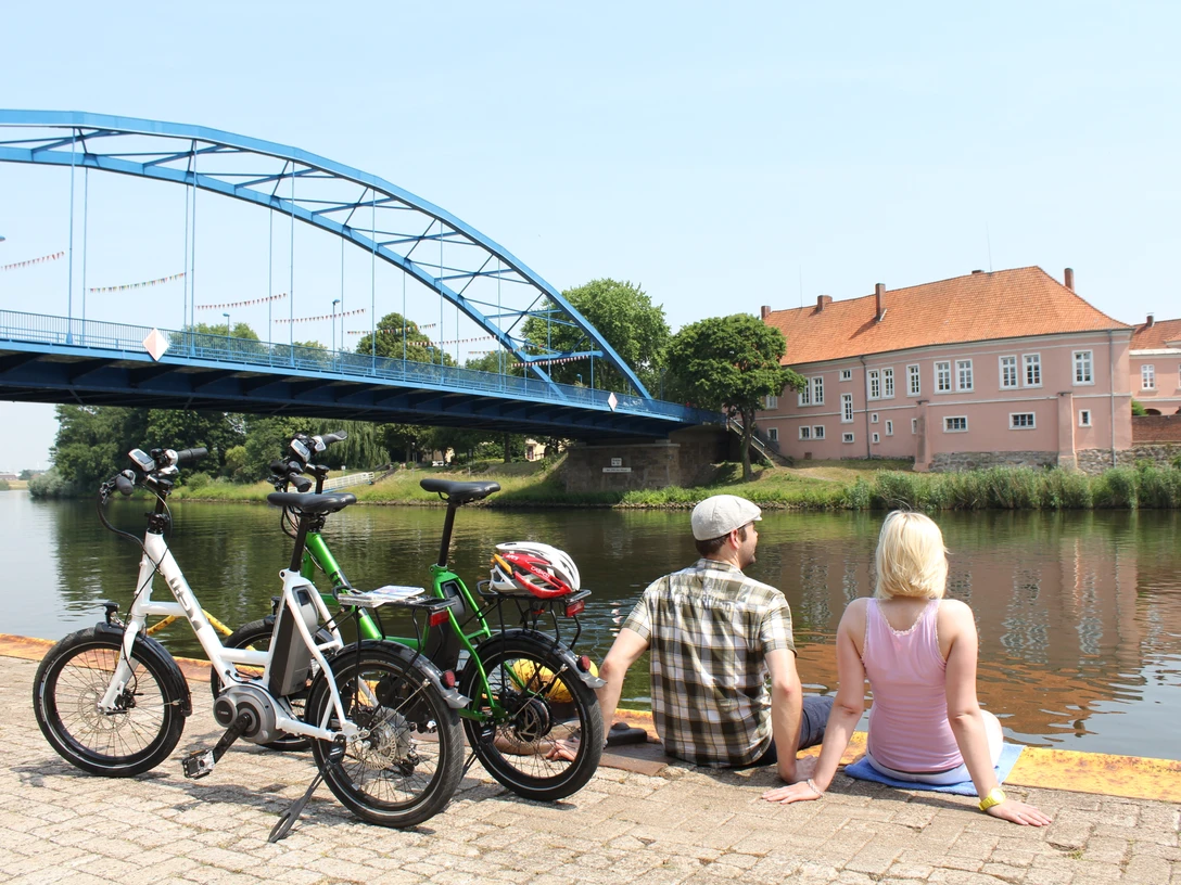Zwei Personen pausieren an einem Flussufer, neben ihnen stehen Fahrräder, im Hintergrund eine Brücke.