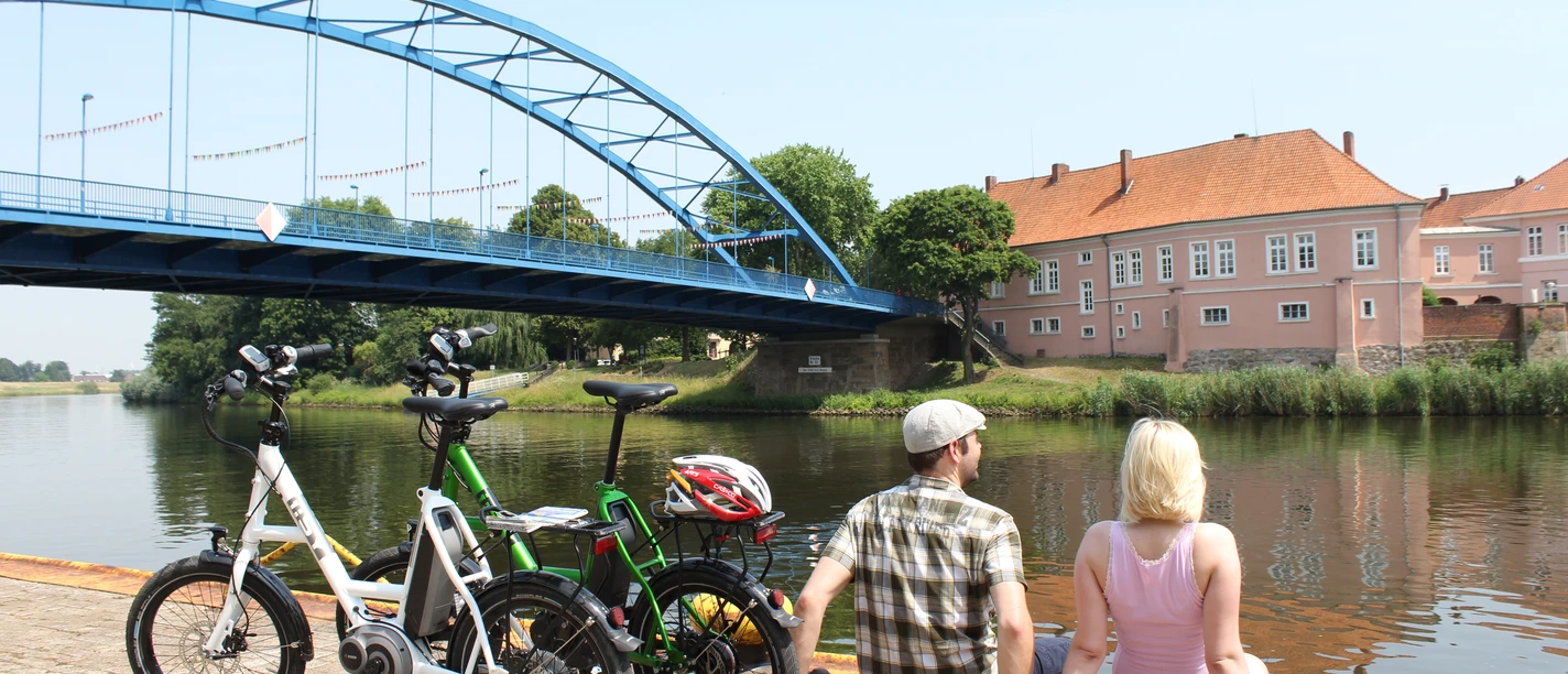 Zwei Personen pausieren an einem Flussufer, neben ihnen stehen Fahrräder, im Hintergrund eine Brücke.