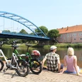 Mittelweser-Region-Radfahren Zwei Personen pausieren an einem Flussufer, neben ihnen stehen Fahrräder, im Hintergrund eine Brücke.