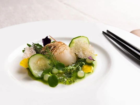 Taku Eine elegante Jakobsmuschel ruht auf einem weißen Teller, umgeben von filigranen Kräutern, Gurkenscheiben und Schaum, serviert mit Essstäbchen.An elegant scallop placed on a white plate, surrounded by delicate herbs, cucumber slices, and foam, served alongside chopsticks.