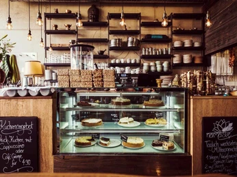 Masala Empire Darstellung eines gemütlichen Restaurants mit einer warmen Atmosphäre. Vorne ist eine Vitrine mit einer Auswahl an Kuchen und Torten zu sehen, die liebevoll angerichtet sind. Die Dekoration besteht aus Holzelementen und hängenden Glühbirnen. Links und rechts neben der Vitrine sind Tafeln mit Kreidebeschriftungen angebracht, die auf Tagesangebote und Spezialitäten hinweisen. Depiction of a cosy restaurant with a warm atmosphere. At the front, there is a display case with a selection of cakes and tarts, lovingly arranged. The décor consists of wooden elements and hanging light bulbs. To the left and right of the display case are chalkboards with chalk writing indicating the daily specials and specialities.