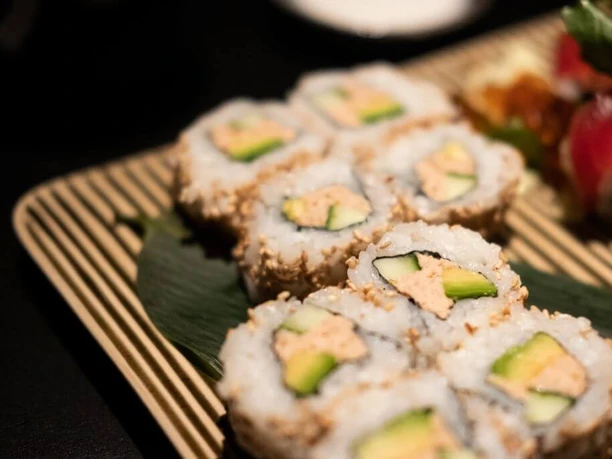 Zen Das Bild zeigt köstlich angerichtete Sushi-Rollen auf einem Bambusbrett, garniert mit Lachs, Avocado und Sesam.