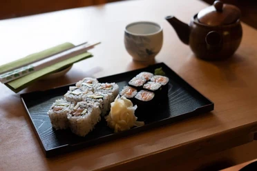 Fisch Hof Sushi Haus Sushi präsentiert auf einem schwarzen Teller. Im Hintergrund eine Kupferteekanne.