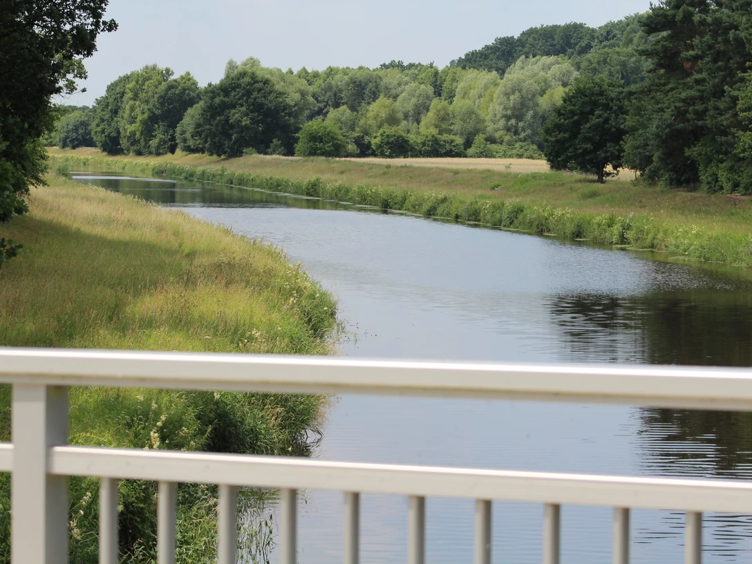 Grüne Landschaft mit ruhigem Fluss, gesäumt von Bäumen, gesehen von einer weißen Brücke aus.