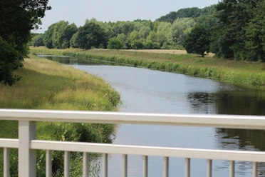 Große Aue Grüne Landschaft mit ruhigem Fluss, gesäumt von Bäumen, gesehen von einer weißen Brücke aus.