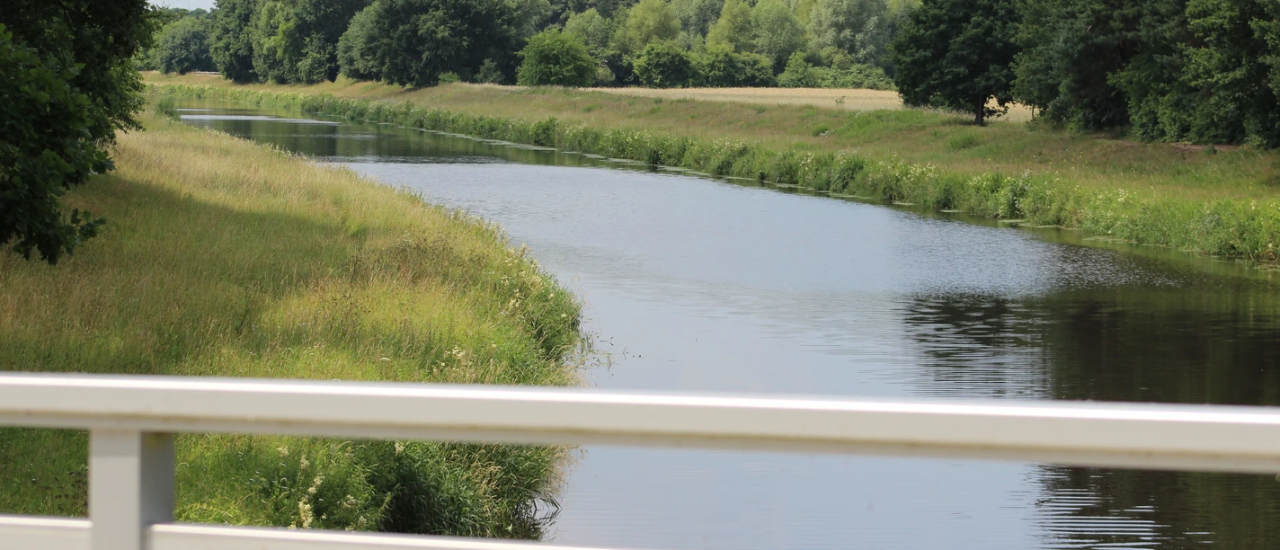 Große Aue Grüne Landschaft mit ruhigem Fluss, gesäumt von Bäumen, gesehen von einer weißen Brücke aus.
