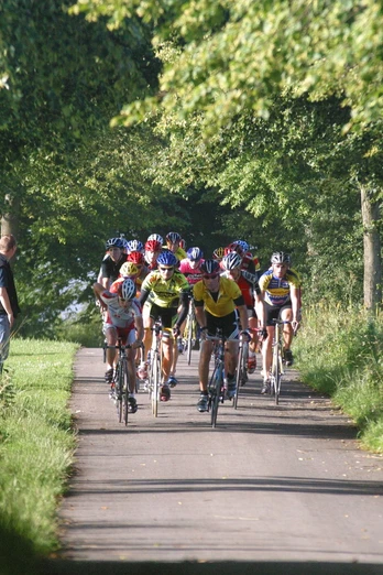 Eine Radfahrergruppe fährt auf einem schmalen, von Bäumen gesäumten Weg durch eine grüne Landschaft.