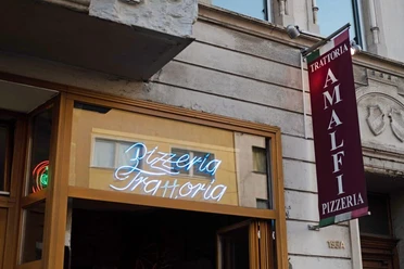 Trattoria Amalfi Außenansicht der "Trattoria Amalfi Pizzeria" mit einem auffälligen Leuchtschild und einem vertikalen Banner an der Wand.