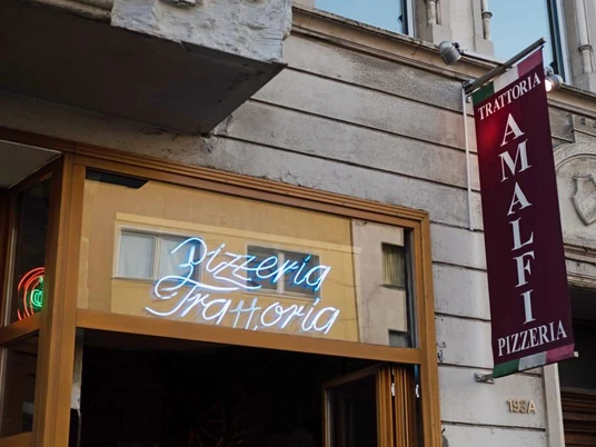 Trattoria Amalfi Außenansicht der "Trattoria Amalfi Pizzeria" mit einem auffälligen Leuchtschild und einem vertikalen Banner an der Wand.Exterior view of "Trattoria Amalfi Pizzeria" featuring a striking illuminated sign and a vertical banner attached to the wall.