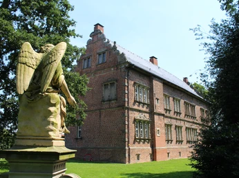 Backstein-Schloss Erbhof in Thedinghausen mit Engelsstatue im Vorgarten, umgeben von grünen Bäumen.