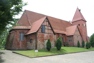 Backsteinkirche mit mittelalterlichem Fachwerk und Spitzdach, umgeben von Grün und blauem Himmel.