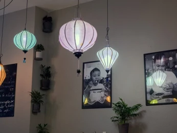 Nam La Vietnamienne Cuisine Eine Wand mit mehreren bunten Papierlaternen, die von der Decke hängen. Daneben sind Schwarz-Weiß-Fotografien zu sehen, die vietnamesische Motive darstellen. Im Vordergrund stehen Pflanzen, die zu einer gemütlichen Atmosphäre beitragen.A wall with several colourful paper lanterns hanging from the ceiling. Next to them are black-and-white photographs depicting Vietnamese motifs. In the foreground are plants that contribute to the cosy atmosphere.
