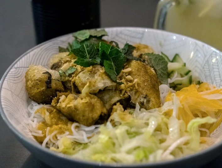 Nam La Vietnamienne Cuisine Eine kunstvoll arrangierte vietnamesische Schale mit Reisnudeln, knusprig gebratenem Tofu und frischen Kräutern auf Reisnudeln, garniert mit knackigem Gemüse.An artfully arranged Vietnamese bowl with rice noodles, crispy fried tofu and fresh herbs on rice noodles, garnished with crunchy vegetables.