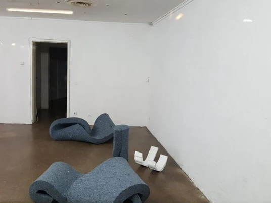 GOLD + BETON Ein leerer, weißer Raum mit geschwungenen dunkelgrauen Skulpturen und einer roten Säule.An empty, white room with curved dark gray sculptures and a red column.