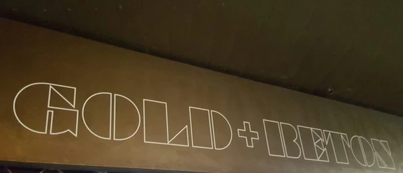 GOLD + BETON Das Bild zeigt eine Wand mit dem Schriftzug "GOLD + BETON" in weiß auf einem gold-braunen Hintergrund.