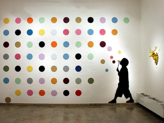 30works Galerie In einem weißen Raum ist eine Installation zu sehen, die aus einer Wand mit bunten Punkten besteht. Daneben die Silhouette eines Kindes, das Blasen pustet, und eine goldene Hirschkopfskulptur.In a white room, there is an installation featuring a wall covered in colorful dots. Next to it is the silhouette of a child blowing bubbles, and a golden deer head sculpture.