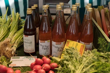 There are various bottles of rhubarb and apple juice on a market stall. Next to them are fresh vegetables such as spring onions, radishes and beet.