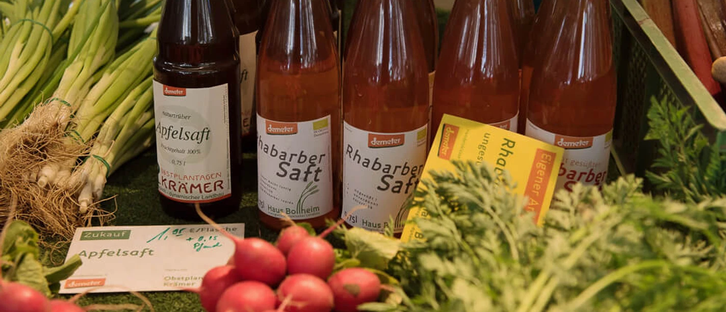 wochenmaerkte_koeln_rudolfplatz-2.jpg Auf einem Marktstand stehen verschiedene Flaschen Rhabarber- und Apfelsaft. Daneben frisches Gemüse wie Lauchzwiebeln, Radieschen und Rote Beete.
