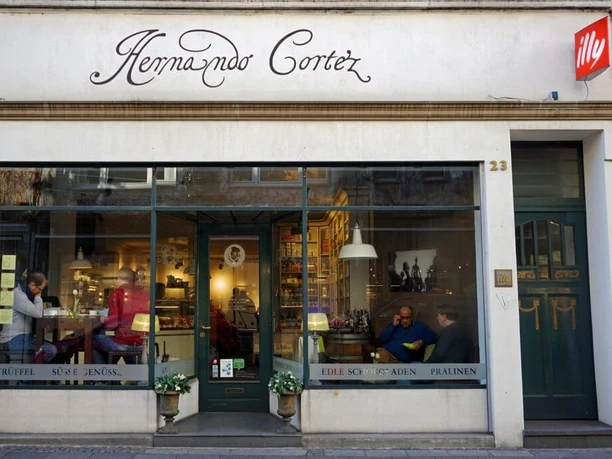 Hernando Cortez Chocolates The façade of "Hernando Cortez Schokoladen" features large shop windows that create an inviting atmosphere. Inside, people enjoy the pleasure of fine chocolate while the stylish lettering logo is emblazoned above the entrance.