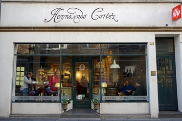 Hernando Cortez Schokoladen Die Fassade der „Hernando Cortez Schokoladen“ zeigt große Schaufenster, die eine einladende Atmosphäre schaffen. Drinnen genießen Menschen den Genuss von edler Schokolade, während das stilvolle Schriftlogo über dem Eingang prangt.