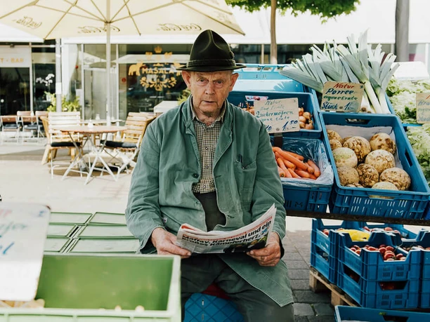 Wochenmarkt auf dem Neptunplatz Ein älterer, traditionell gekleideter Mann sitzt auf einem Marktstand, umgeben von frischem Gemüse.