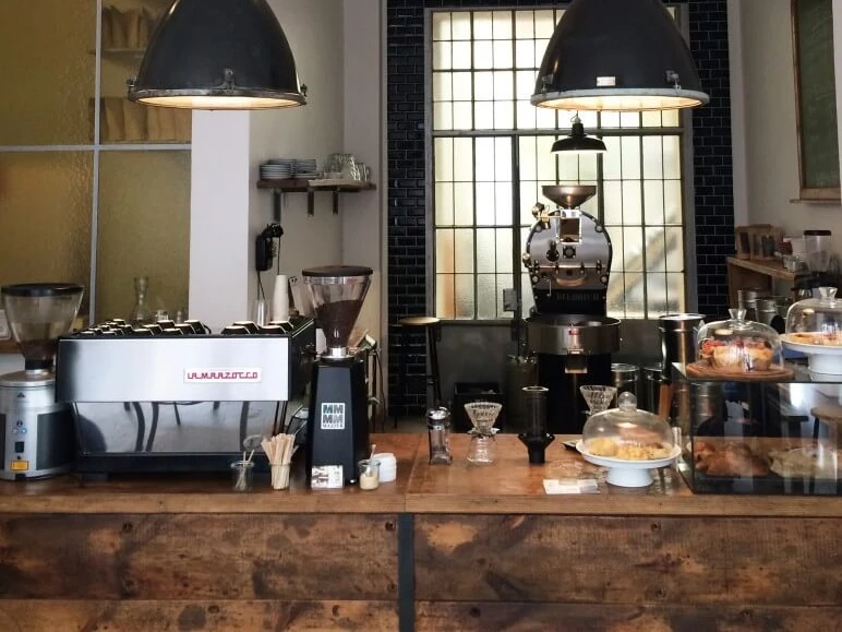 ERNST KAFFERÖSTER Im Industriestil eingerichtete Theke mit Kuchenauslage und Espressomaschine. An industrial-style counter with a cake display and an espresso machine.