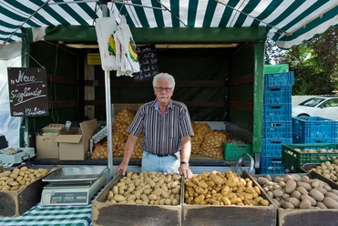 wochenmaerkte_koeln_weiden-3.jpg Älterer Herr verkauft frische Kartoffeln an einem Marktstand unter einem gestreiften Zelt.