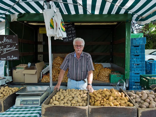 wochenmaerkte_koeln_weiden-3.jpg Älterer Herr verkauft frische Kartoffeln an einem Marktstand unter einem gestreiften Zelt.Elderly man selling fresh potatoes at a market stall under a striped tent.